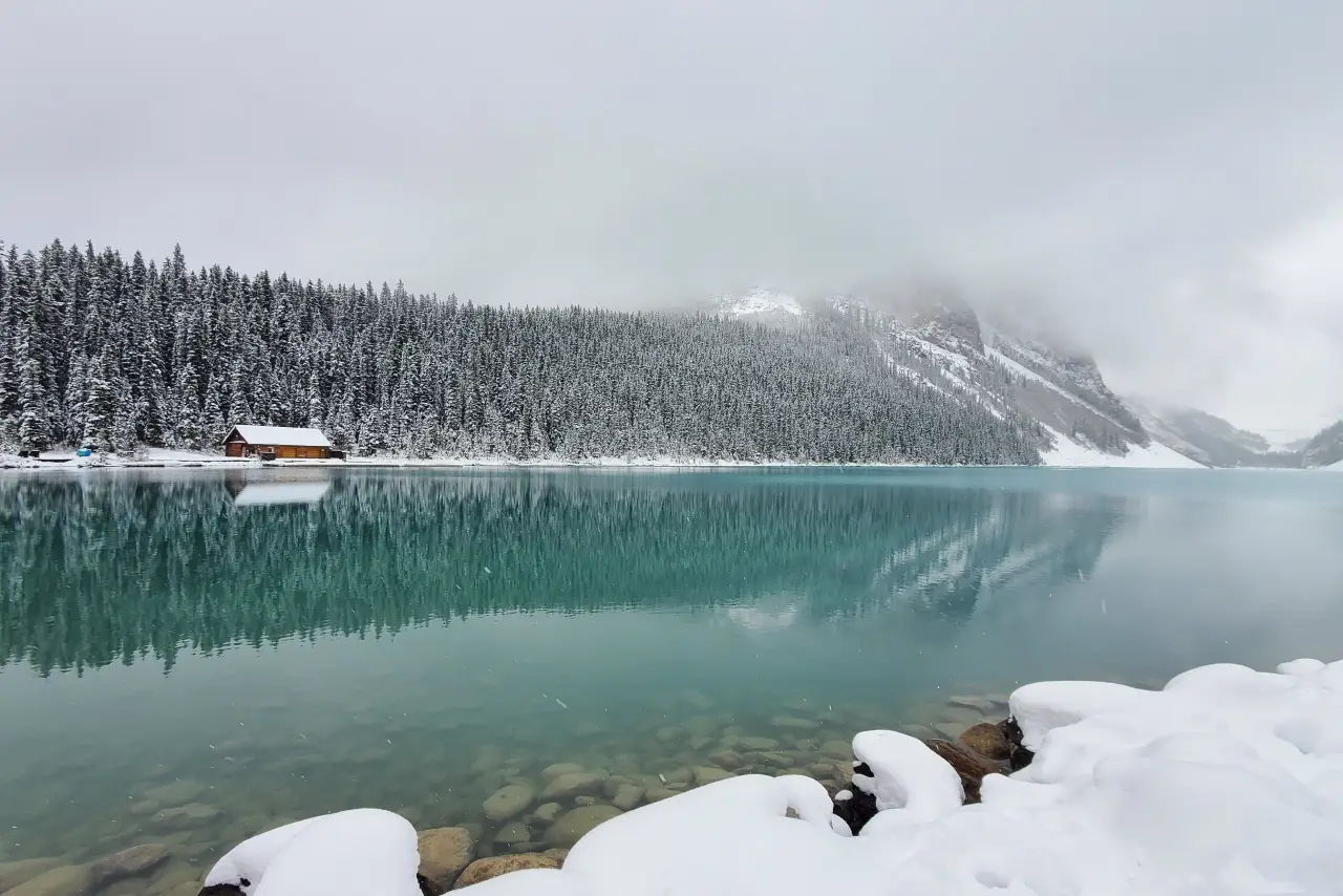 A serene winter scene of a turquoise lake with a snow-covered cabin and forest.