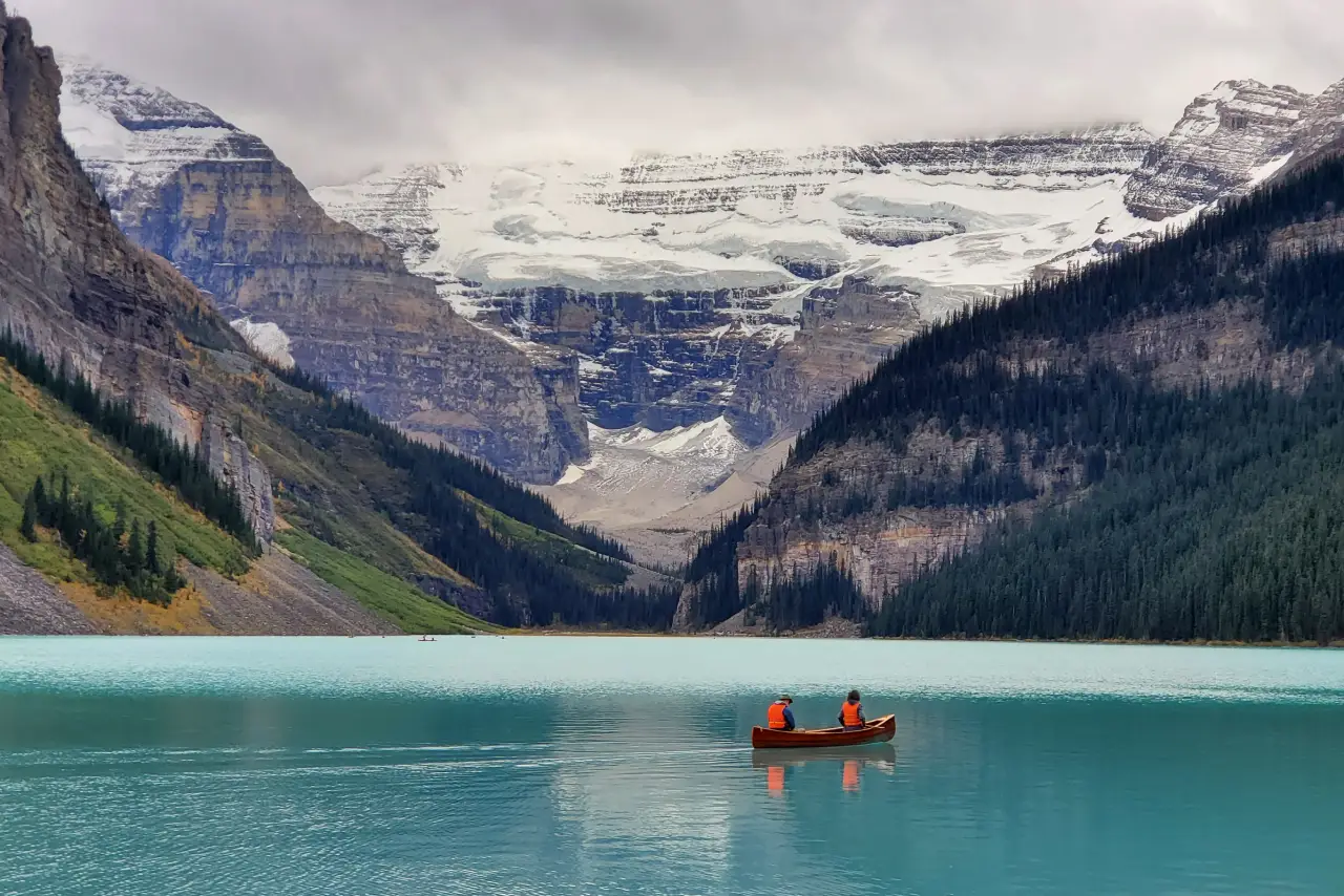 Two people in a canoe on a turquoise lake with snowy mountains in the background.